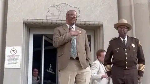 In the image, we see a man standing in front of a building with his hand over his chest. He is looking out to the people. Next to him, we see a sheriff.