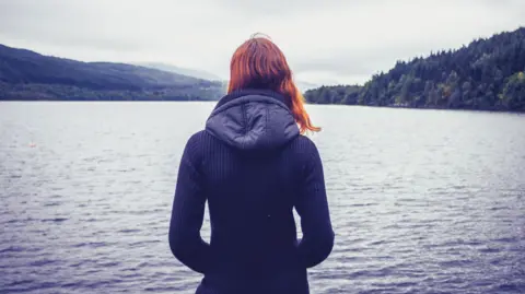 Young woman with her back to the camera looking out over a misty Scottish loch - generic. She has red hair and is wearing a blue jacket with a hood.