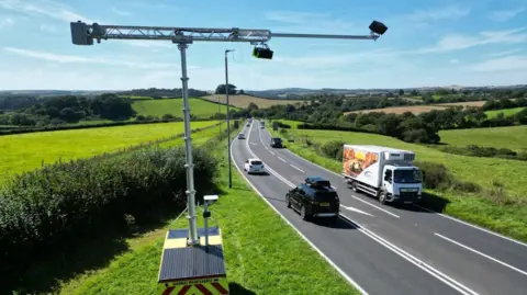 The camera was rolled out on the A38 in 2023 - you can see the stretch of road with the camera on grassland next to it. It is a small trailer with a large pole running upwards and over the top of the carriageway.