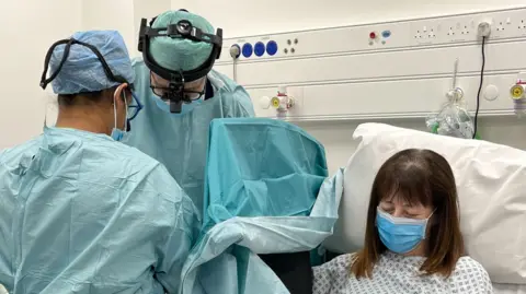 A female patient in a hospital gown with a blue face mask on sits in a chair. To the left of her, two surgeons dressed in blue surgical clothes are working on her hand, which is not visible and is behind a screen