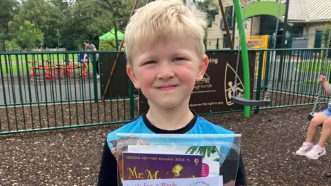 Theo, a boy with blonde hair and a blue shirt holds up his books