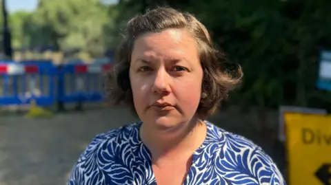 Steph Nissen with ear-length brown hair  and brown eyes looking off into the distance with an unimpressed look on her face. She is wearing a white top with navy leaf swirls on it. The background is blurred but you can make out the closed section of the bridge and a yellow diversion sign to the right