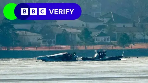 A boat approaches a wrecked helicopter in a river in front of houses