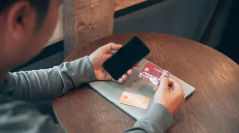 Man holds a credit card and a phone with another bank card and a laptop on the table in front of him.