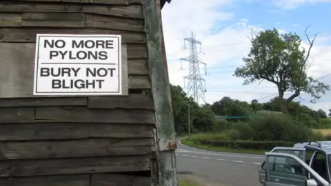 Andrew Woodger/BBC  Sign saying "No more pylons, bury not blight" with a pylon in the background