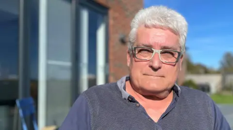 Mike Crookes sits in a garden outside a house on a sunny day. He has bushy, white hair and wears glasses with black and white frames, and a grey and blue polo shirt.