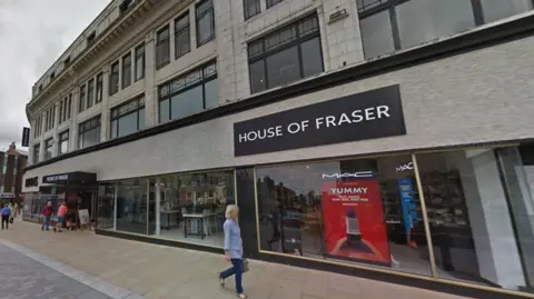 House of Fraser in Darlington. It is a grey three-storey building. There are make-up brands in the shop front. A woman wearing a blue shirt and jeans in passing by. A couple of people are entering the store. 