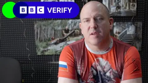 X A bald man looking at the camera wearing a t-shirt with a wolf on the front and a Russian flag on the sleeve