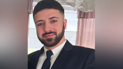 Hertfordshire Police  Kyle Clifford with dark hair and beard wearing a black jacket, white shirt and black tie