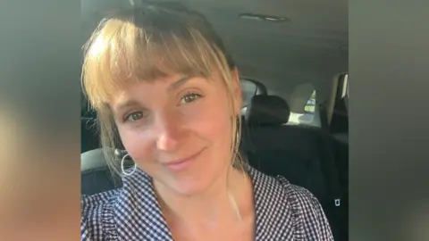 A selfie of Chloe Davies, who has blonde hair and brown eyes. She is sitting in a car and has her head slightly tilted as she looks into the camera and smiles