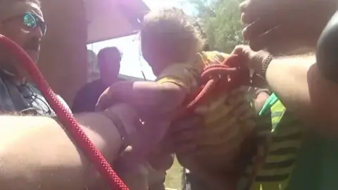 A toddler is surrounded by men helping him get the rope out of him after being rescued.