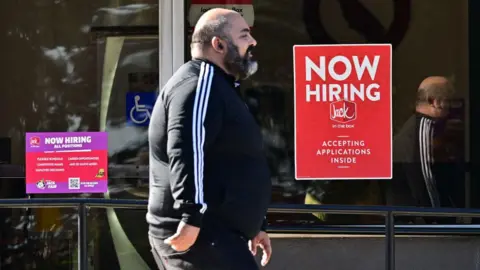 A pedestrian walks past a "Now Hiring" sign posted on a business storefront in San Gabriel, California on August 21, 2024. 