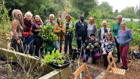 Men and women in a garden in Brierley Hill stand in a row together in the photo. They have plants in front of them and behind and two women are holding plants in their hands.