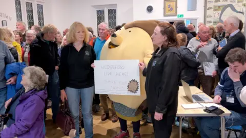 Campaigners in a packed village hall, one of whom is dressed as a hedgehog. Placard says "Hedgehogs live here!"