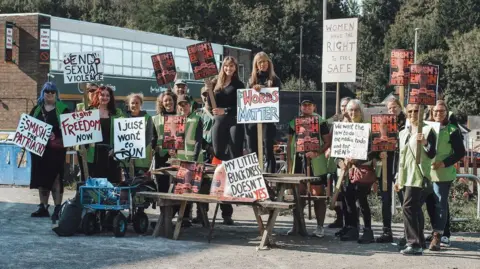 A group of women are sitting and standing on a bench holding placards which read 'My little black dress doesn't mean yes' and 'Women have the right to feel safe.'