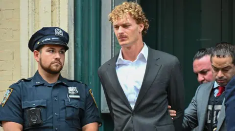 Daniel Penny in grey suit with no tie with his arms behind his back, next to police officer in uniform and being escorted by police officer in suit jacket.