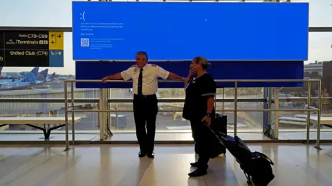 United Airlines employees wait by a departures monitor displaying a blue error screen.