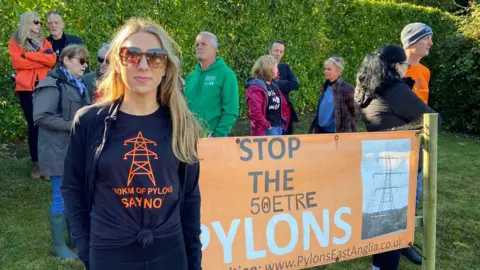 A photo of Nicole Gibson. She is facing the camera and standing in front of other protesters. She is standing in front of a sign that reads 'stop the pylons'. She has blonde hair and is wearing sunglasses. She is wearing a black t-shirt that reads '180km of pylons, say no'. She is also wearing a black jacket.