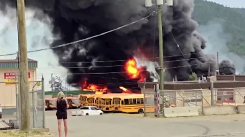 A woman stands in front of a raging fire and a huge plume of black smoke rising above yellow school buses