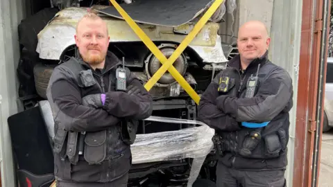 PC Paul Gerrish and PC Phil Pentelow are wearing grey jackets and trousers and have radios on their lapels. They are standing with their arms folded in front of stolen cars with yellow tape across the vehicles,