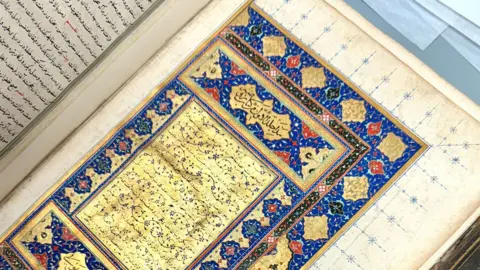 An open 16th Century book showing a writing on gold edged with bright blue, red, black and pink decorations Rawḍat al-ṣaffāʾ (Garden of Purity) by Muḥammad bin Khāwand Shāh