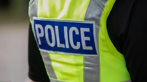 Back of a British police vest that is bright yellow with silver stripes running down the side and a blue rectangle with the word POLICE is bold white lettering.