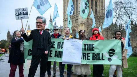 Eight protesters in favour of the bill gather outside Parliament. One is dressed as Keir Starmer and wearing a mask with the face of the prime minister. He is holding a sign reading: "Let the CAN bill live"