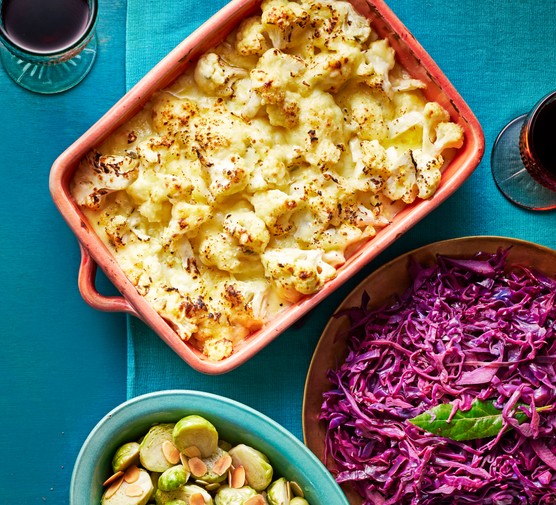 Cauliflower cheese in an orange dish next to brussel sprouts and red cabbage