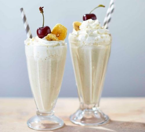 Two glasses of banana milkshake topped with cream and fruit