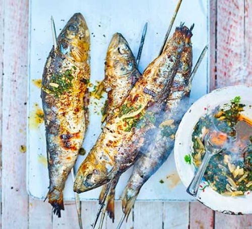 Skewered barbecue sardines on a board by a plate of chermoula sauce