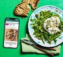 A phone with the Good Food app on its screen plus a green salad topped with burrata