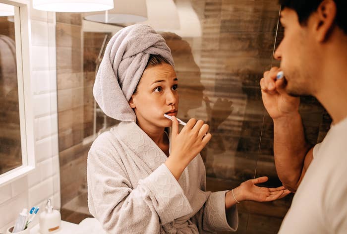 A woman with a towel wrapped around her head and wearing a robe brushes her teeth while talking to a man also brushing his teeth in a bathroom