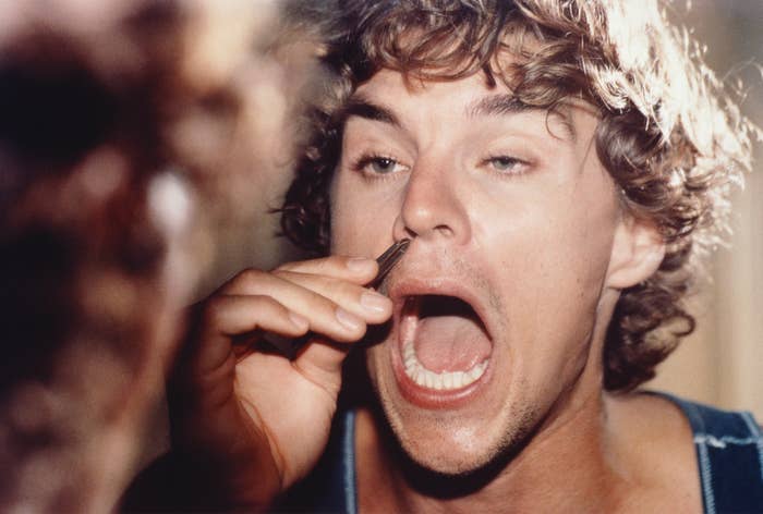 A person with curly hair holds a pair of pliers up to their nose and opens their mouth wide in a dramatic expression