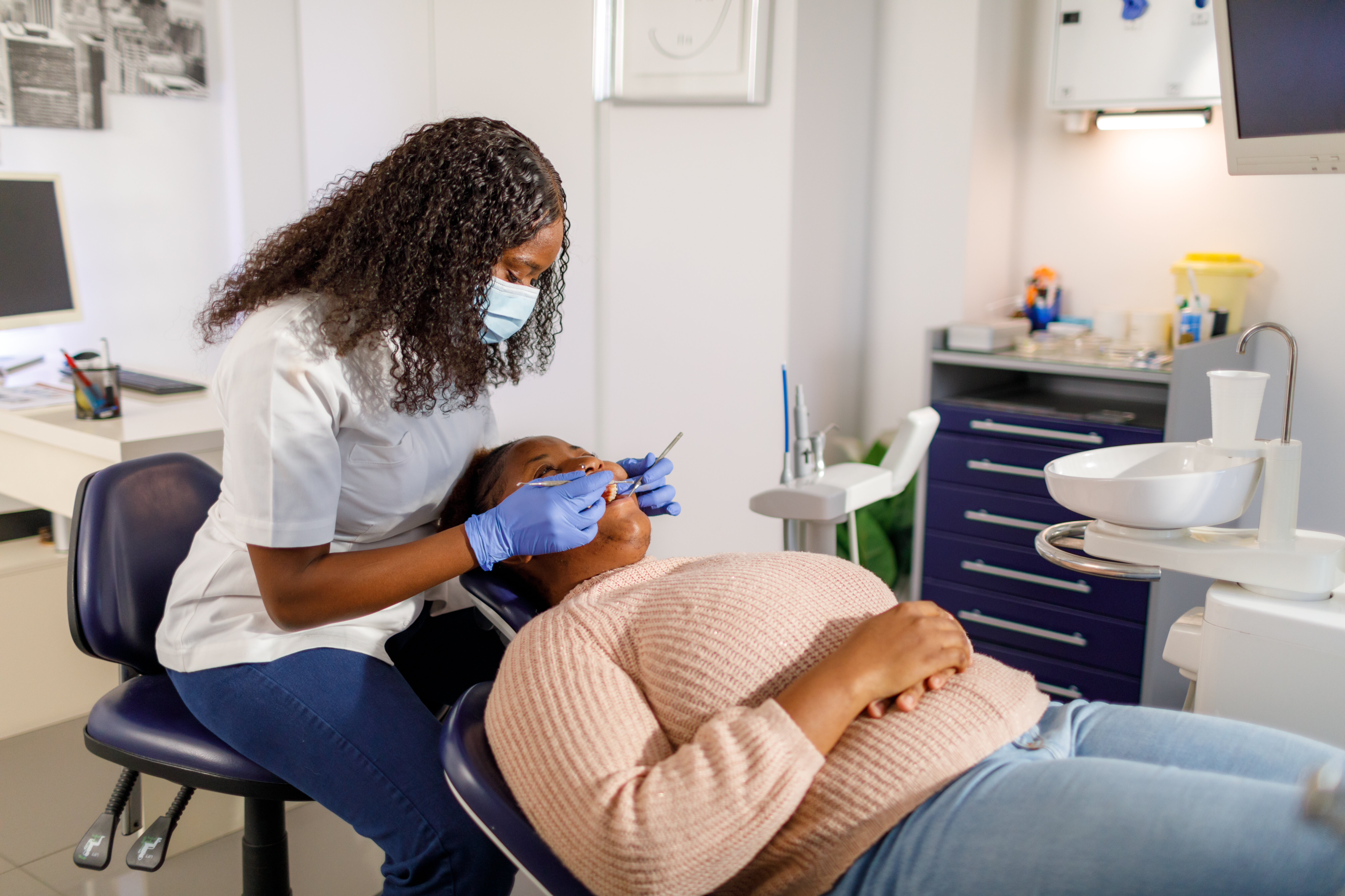 Alicia Keyes, in a pink sweater, is receiving dental care from Dr. Sandra Brown, who is wearing a white coat and face mask, in a modern dental office