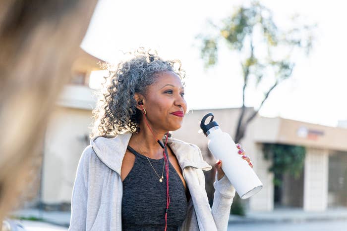 A woman with curly hair, dressed in activewear and earphones, smiles while holding a water bottle during an outdoor workout session