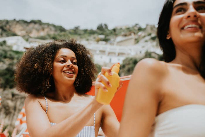 Two women smiling while applying sunscreen to each other on a beach. Houses are visible on the hillside in the background