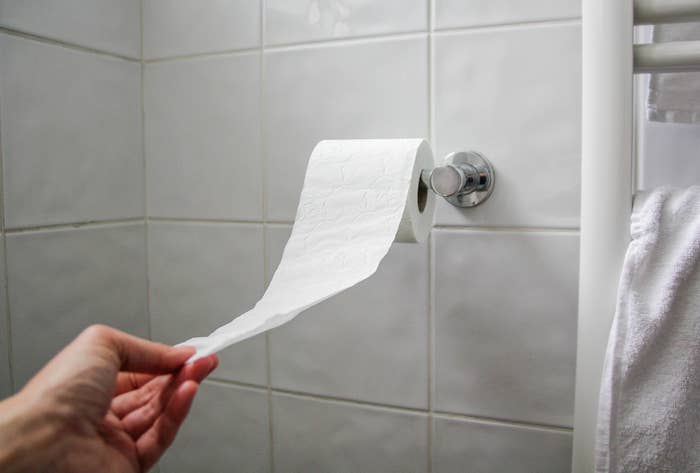 A hand pulling toilet paper from a wall-mounted roll next to a towel rack in a bathroom