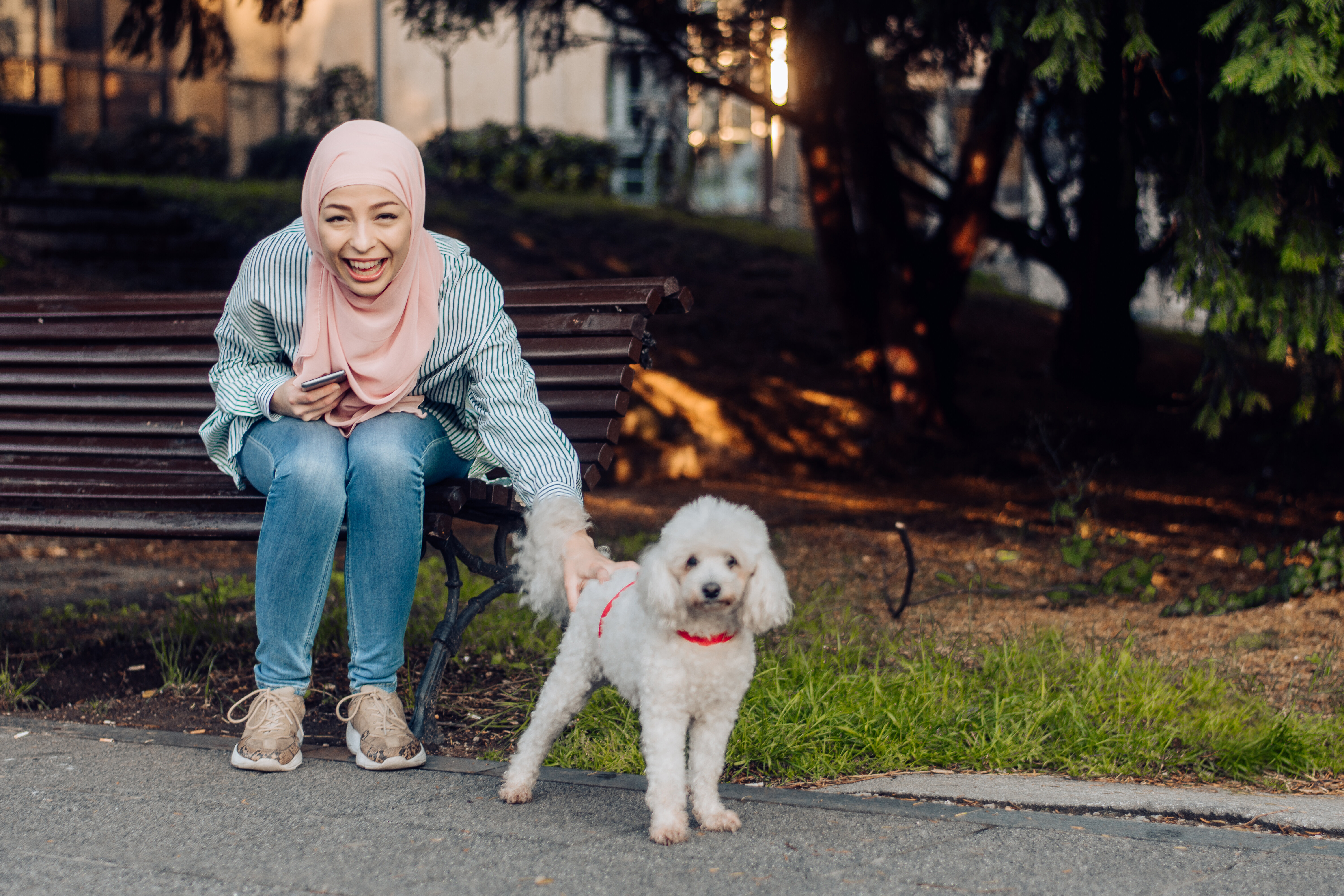 A woman in casual clothing sits on a park bench, smiling and petting a small white dog with a red collar