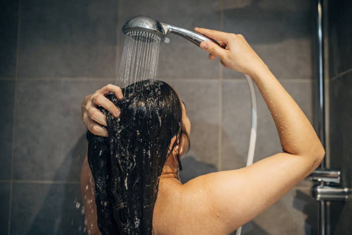 A person showers, holding a showerhead above their head and rinsing their hair
