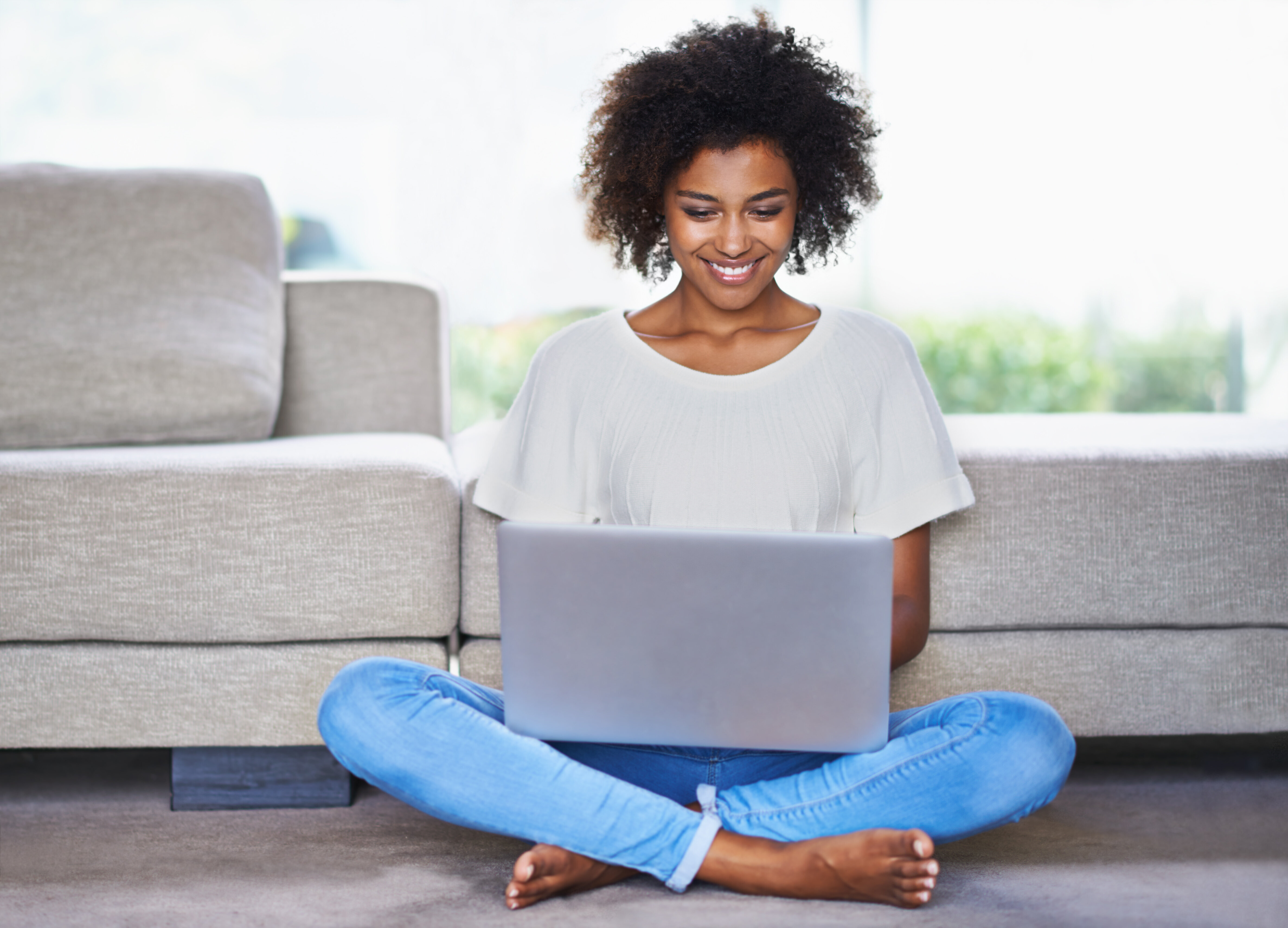 Woman sitting on the floor, legs crossed, smiling while using a laptop in a cozy living room with a couch in the background