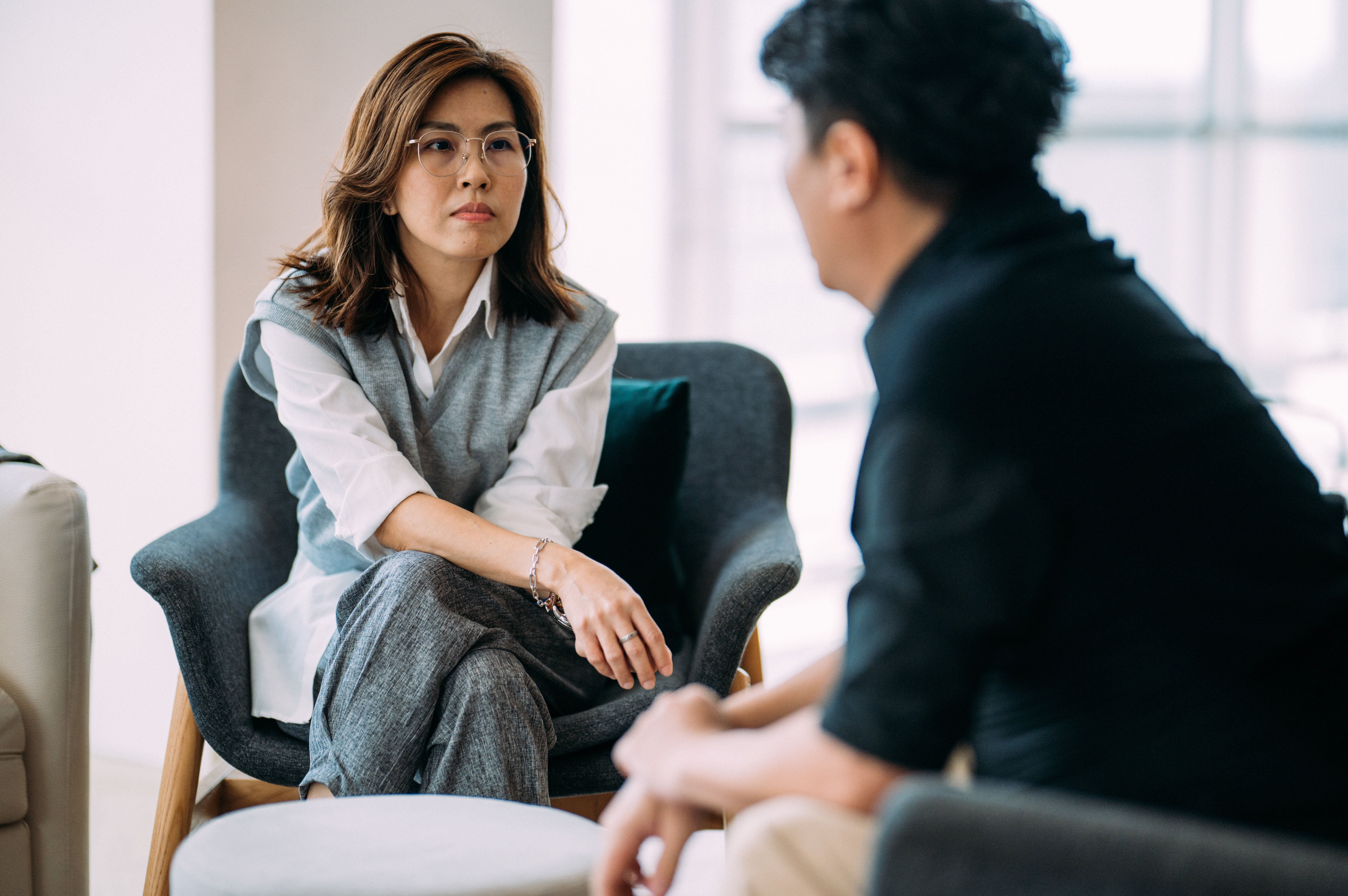 A woman with glasses and a man in conversation in a modern office setting. The woman has a thoughtful expression and is seated with legs crossed