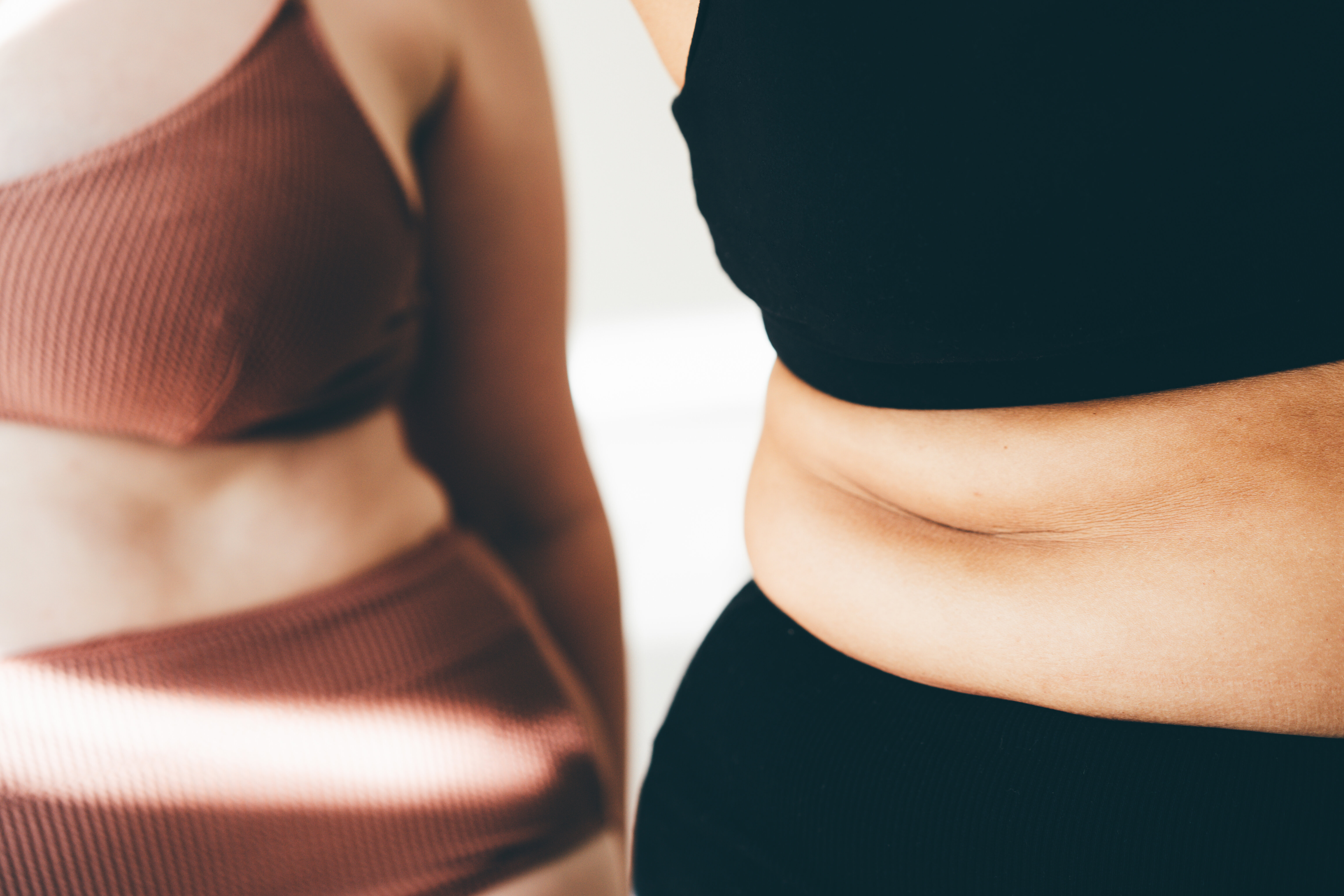 Two women in stylish, ribbed activewear tops and high-waisted bottoms are shown close up, highlighting body positivity and self-love. Names not known