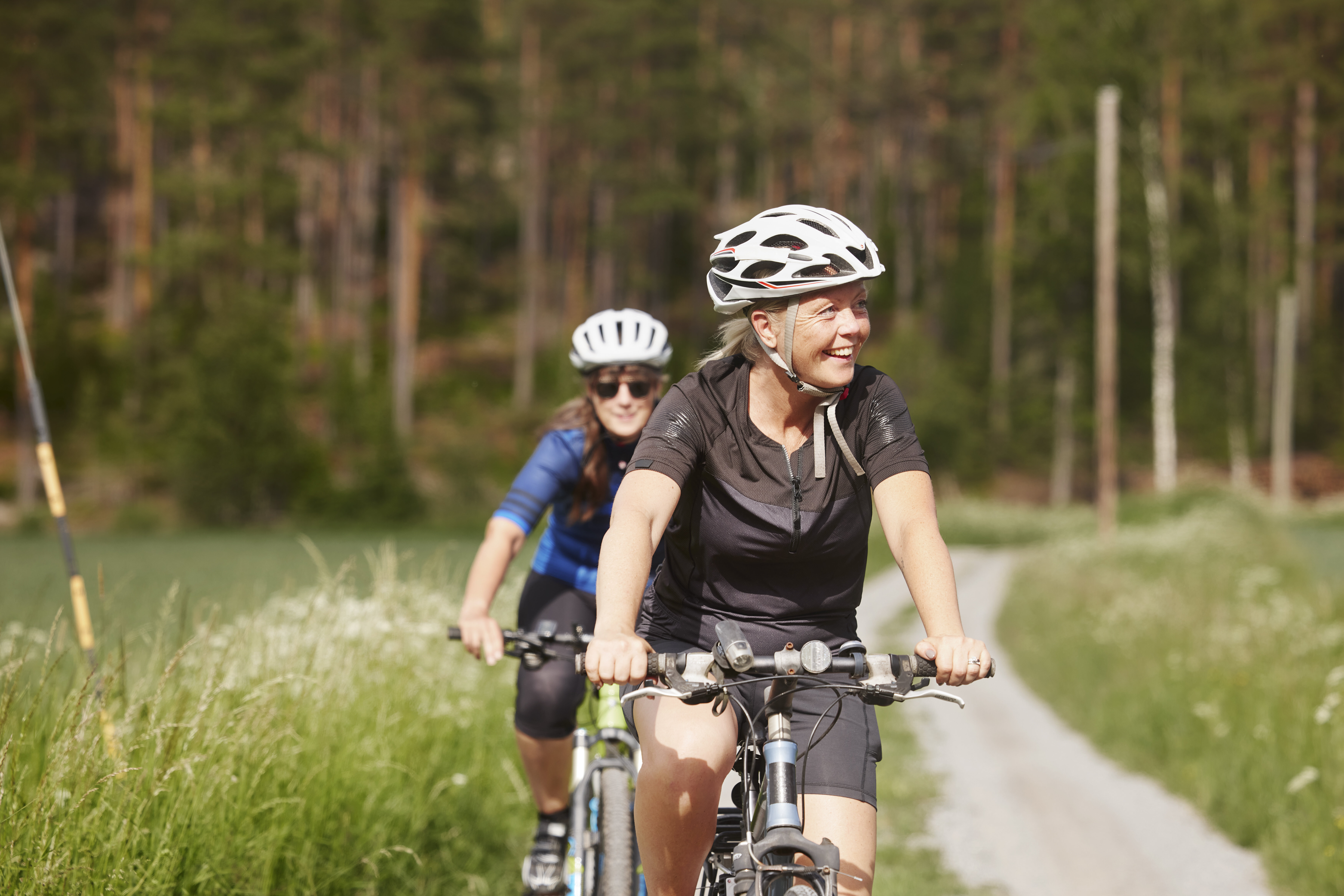 Two women riding bicycles on a countryside path, both wearing helmets and cycling gear, with a forest in the background