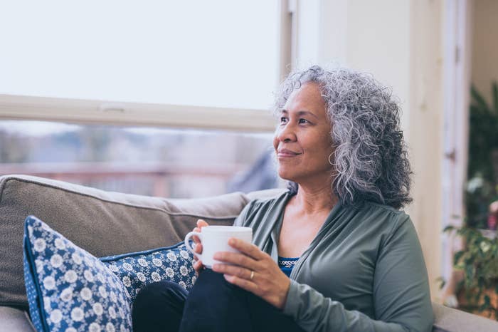 A woman with curly gray hair and a serene expression sits on a couch holding a mug, looking out the window. She wears a casual, relaxed outfit