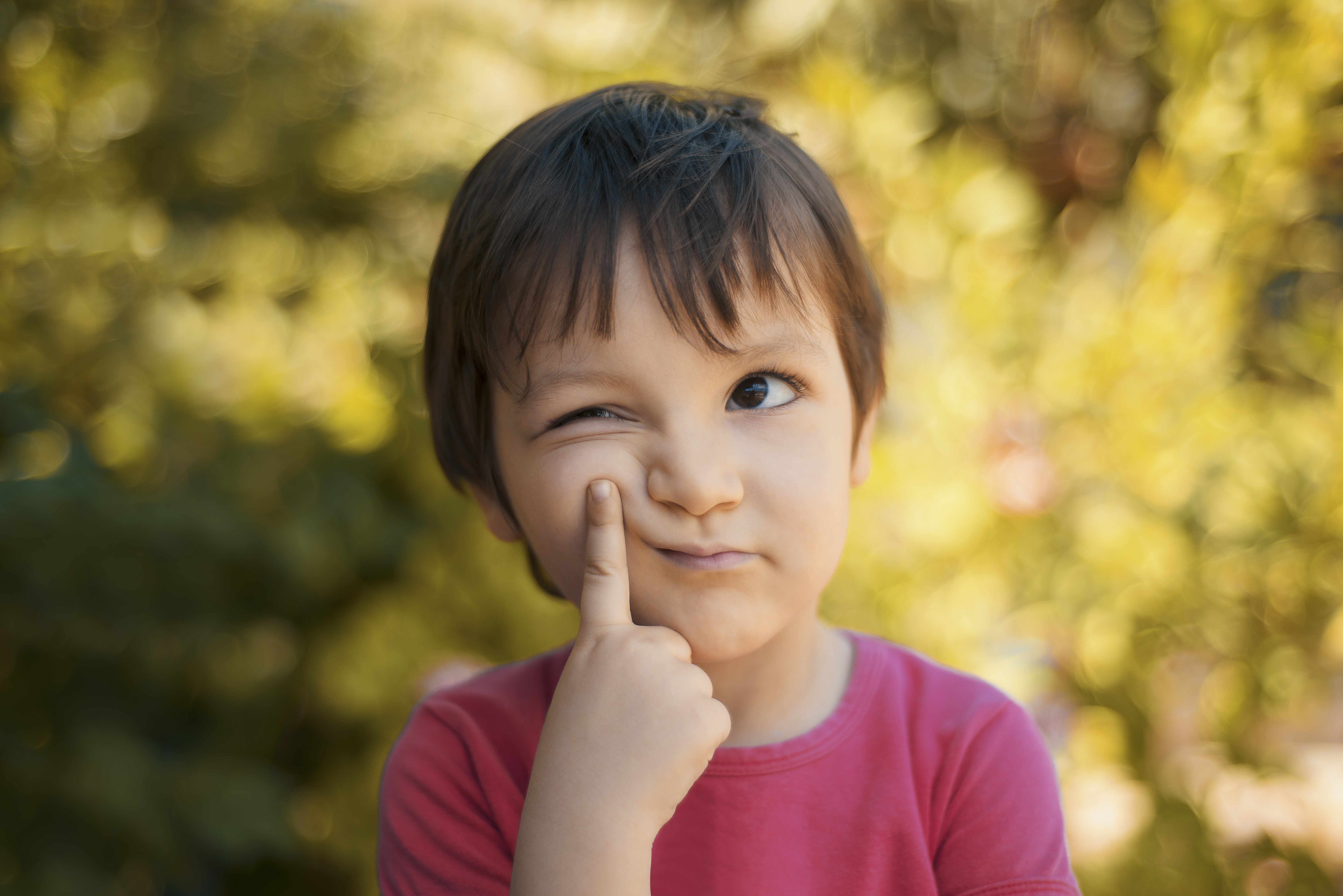 A young child wearing a simple shirt stands outdoors, looking thoughtful and playfully pressing one finger to their cheek. The background is blurred foliage