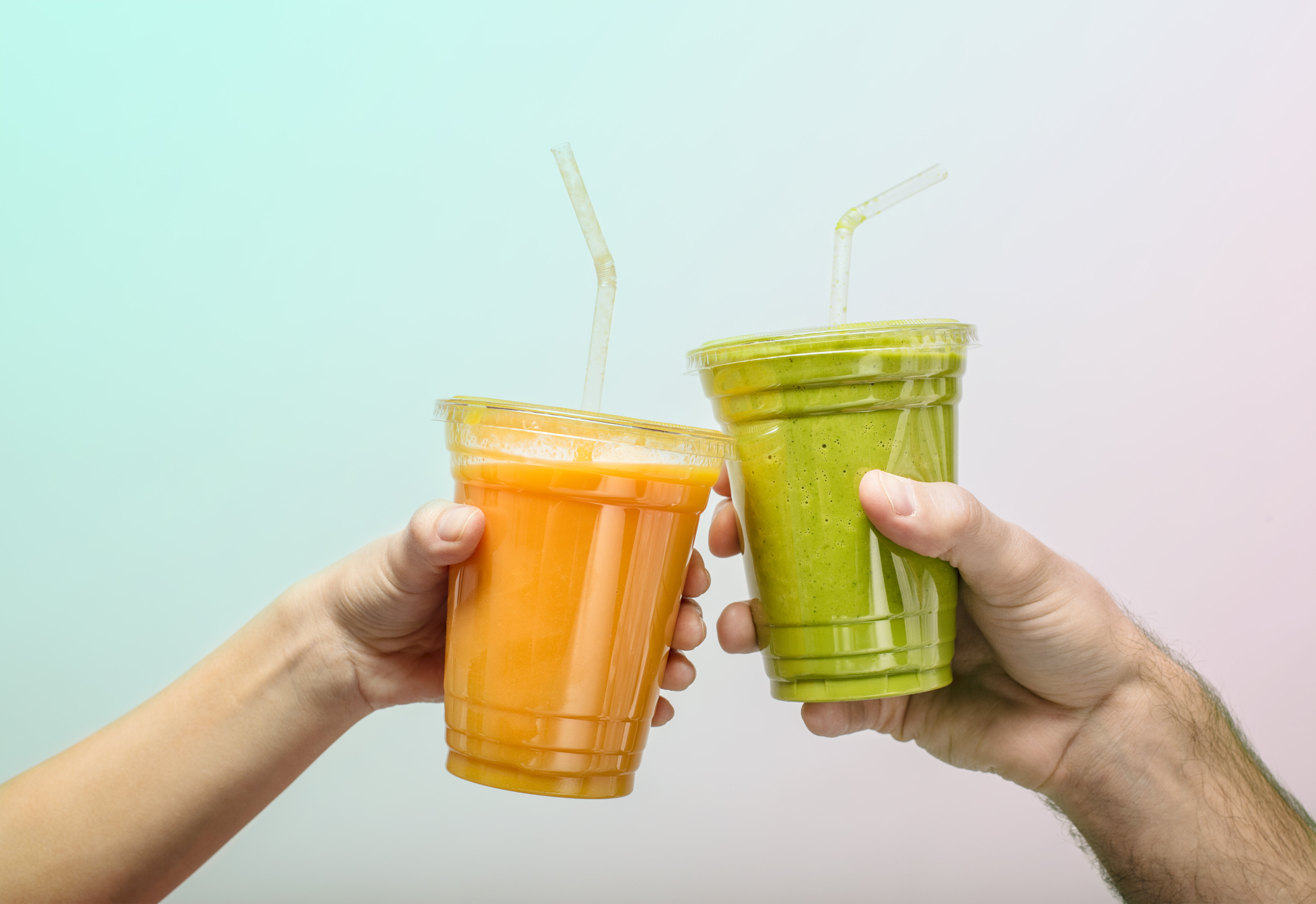 Two hands holding plastic cups with straws, one with an orange drink and the other with a green smoothie, toasting together