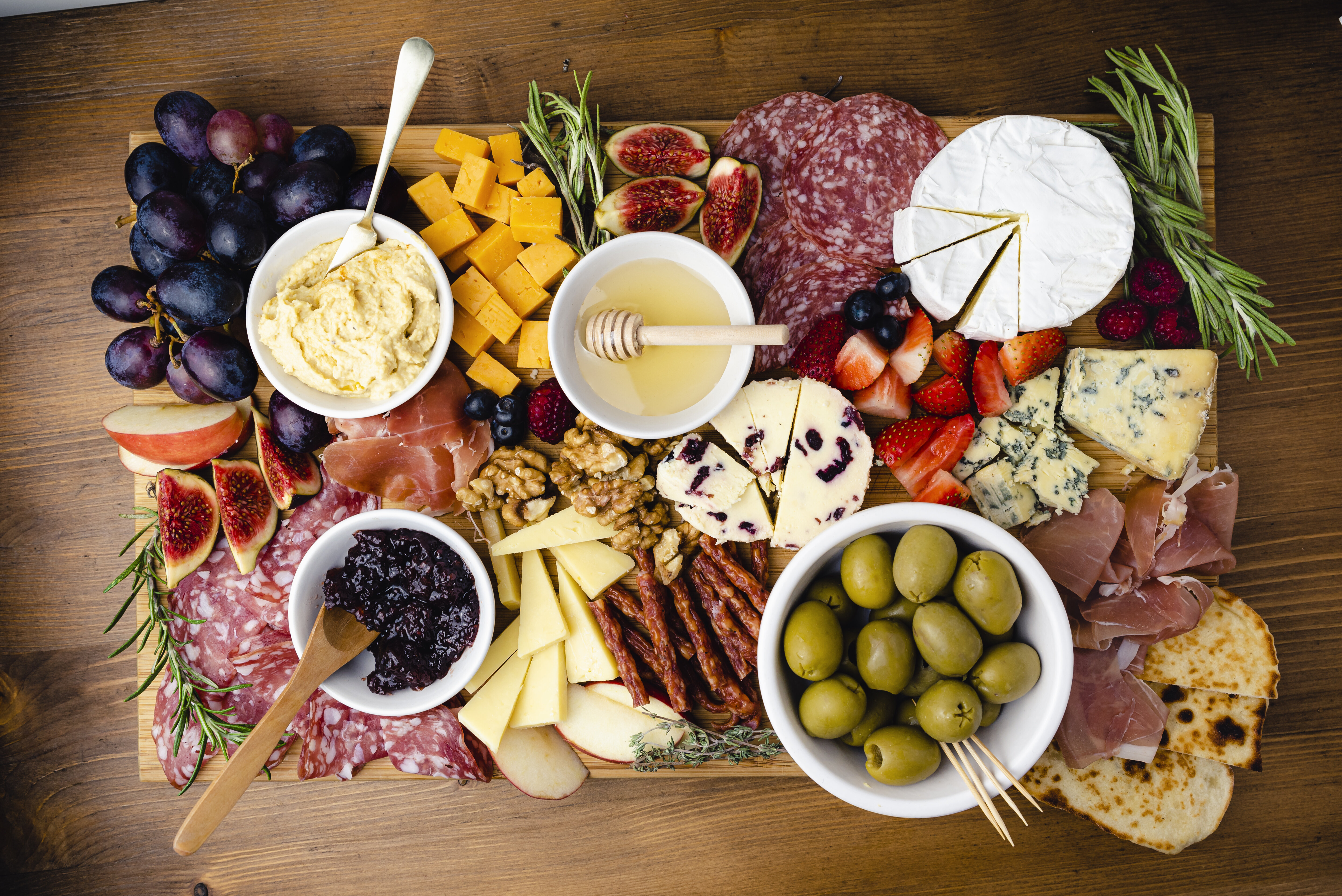 A charcuterie board with various cheeses, meats, fruits, crackers, olives, nuts, honey, and dips. Items are artistically arranged on a wooden board