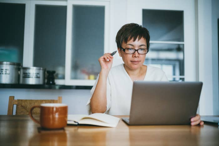 A woman wearing glasses and a white blouse works intently on her laptop at a home office. She is holding a pen and has an open notebook beside her