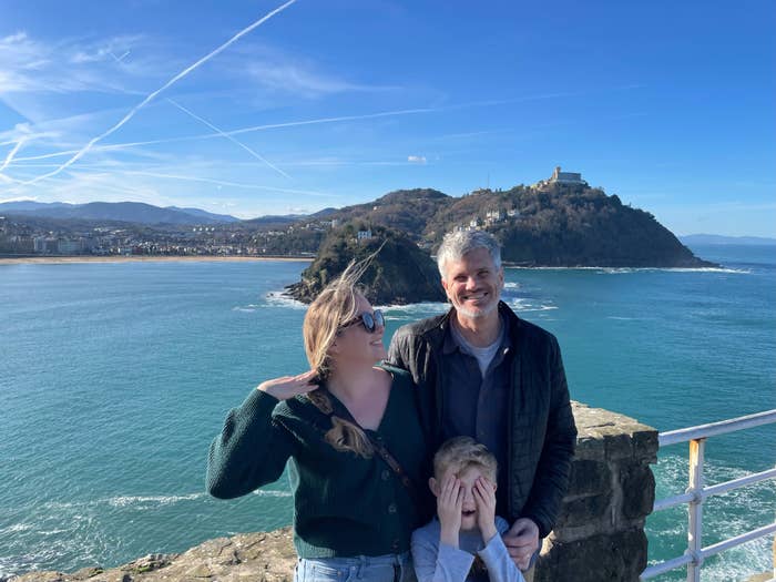 A woman, a man, and a child are posing on a scenic viewpoint with mountains, a large body of water, and an island in the background. The child is covering their face