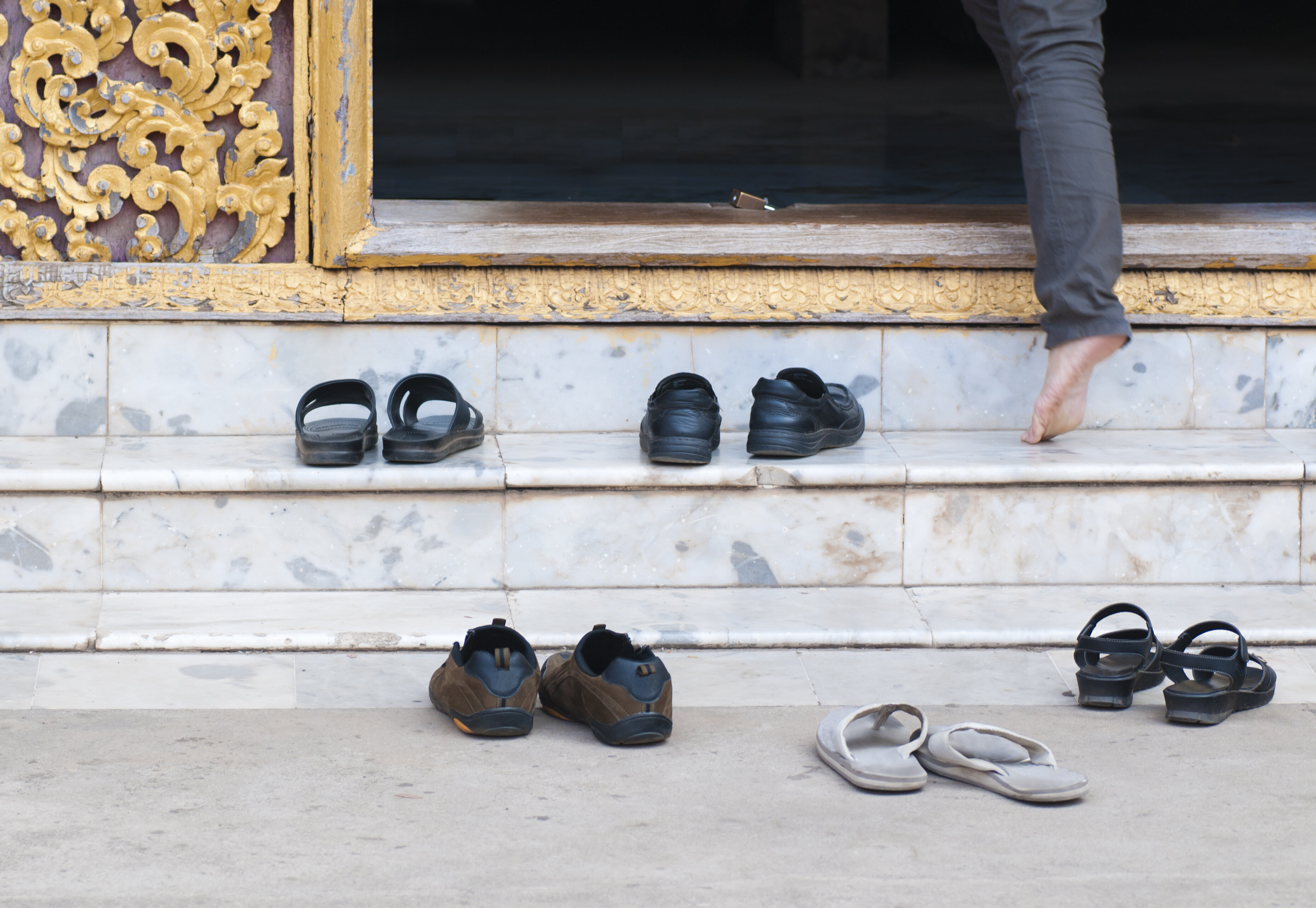 Several pairs of shoes are placed outside on marble steps near an ornate golden door. A person is seen stepping into the building barefoot